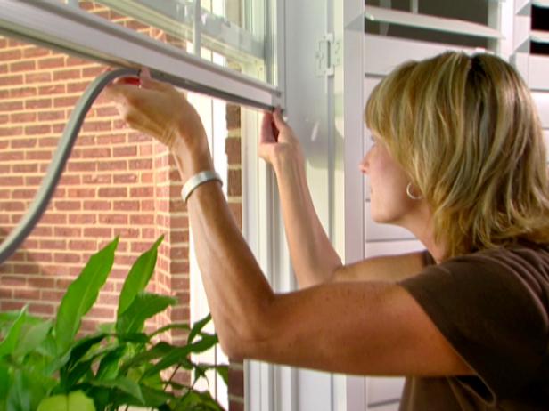 A woman adjusts a window screen indoors, with a plant and a brick wall in the background.