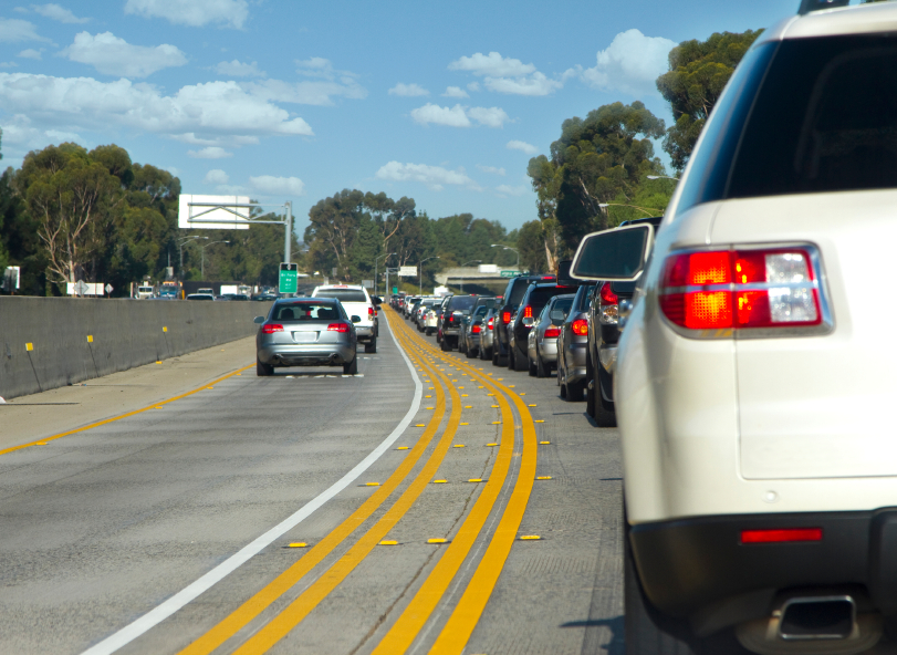 Heavy traffic on a multi-lane highway with cars lined up bumper-to-bumper in two lanes. The sky is clear, with scattered clouds, and trees line the sides of the road. A highway sign is visible in the distance above the road.