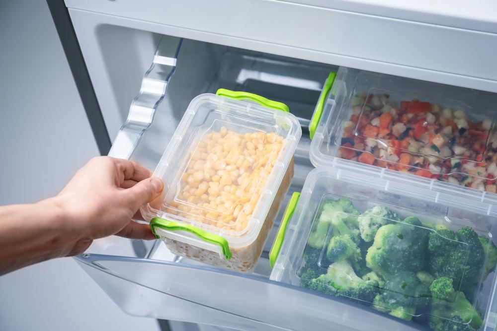 Hand holding a container of frozen corn above a freezer drawer with other containers of frozen vegetables.