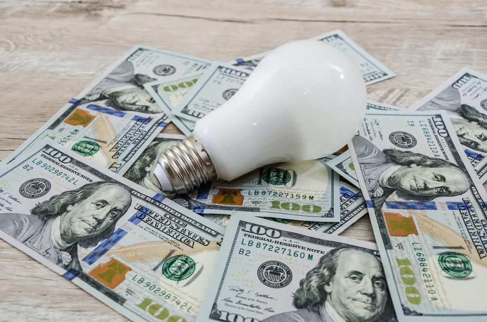 A white light bulb lying on top of scattered 100-dollar bills.