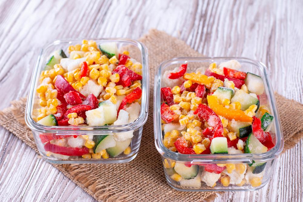 Two glass containers filled with a mix of corn, bell peppers, cucumbers, and cauliflower on a burlap fabric over a wooden surface.