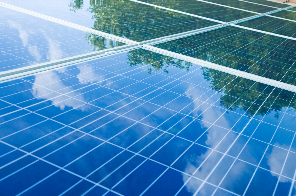  A close-up view of solar panels reflecting the outline of nearby trees and a blue sky with scattered white clouds. The solar panels are aligned in a grid pattern, capturing the reflections from above.