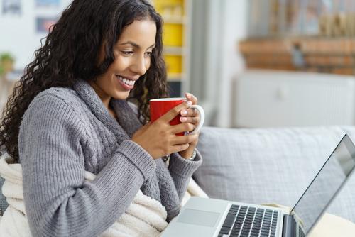 A woman in a gray sweater smiling while holding a red mug and looking at a laptop screen.