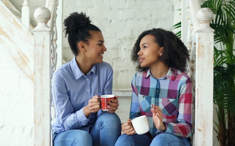  Two women sitting on a rustic, white-painted staircase with an ornamented banister. Both are casually dressed in blue jeans holding mugs and talking.