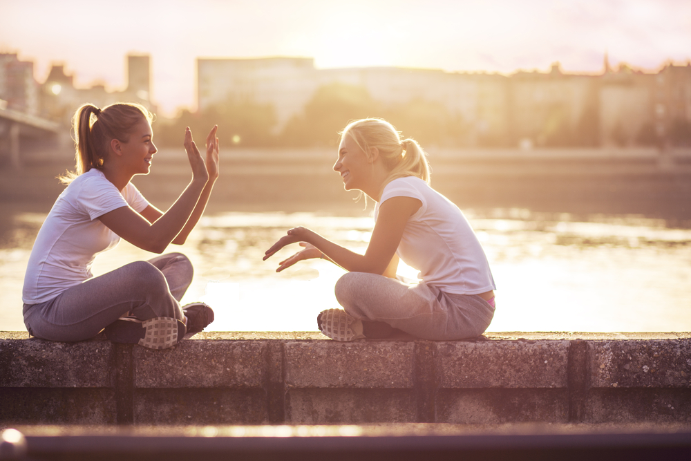  Two women sitting on a low concrete wall, facing each other and interacting playfully at a riverside during sunset. 